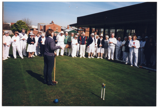 COL photo of a woman wearing blue trousers and jacket, addressing a group of people on a grass croquet court. She is holding a mallet, the head of which is resting on the grass. Beside her, on the grass court, there is a hoop with croquet clips attached and a ball. The people in the group are wearing white uniforms, with either skirts or trousers. A few people are also wearing dark green or dark blue cardigans or jumpers. One man is wearing a dark blue and red track suit jacket. One woman is wearing an 'Active for Life' branded t-shirt. The group is standing on the green, beside a building. In the background, there are several other buildings alongside a wooden fence.