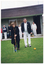 COL photo of a woman posing with a croquet mallet, as if about to hit a ball through a hoop. There is a man standing beside her. They are standing outdoors on a grass croquet court. In the background, there is a building, with several people standing in a row under the eaves. Some of them are holding croquet mallets.