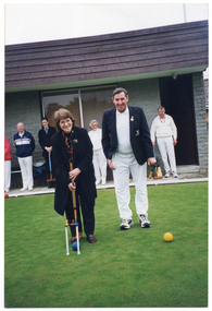 COL photo of a woman posing with a croquet mallet, as if about to hit a ball through a hoop. There is a man standing beside her. They are standing outdoors on a grass croquet court. In the background, there is a building, with several people standing in a row under the eaves. Some of them are holding croquet mallets.