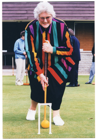 COL photo of a woman posing with a croquet mallet, as if about to hit a ball through a hoop. She is standing outdoors on a grass croquet court. In the background, there are a few other people on the grass court, also holding mallets.