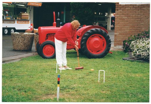 COL photo of a woman holding a mallet, and hitting a ball through a hoop. She is standing on a grassy area. Behind her is a red tractor and a food vendor caravan selling donuts, cappucinos, and drinks. Behind the tractor, there is an undercover carpark and a brick building.