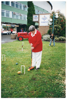 COL photo of a woman holding a mallet, preparing to hit a ball through a hoop. She is standing on a grassy area. Another ball, which is travelling through the air, is about to go through the hoop. In the background, there is another woman holding a mallet - she is crouching forward. There is also a red tractor and a food vendor caravan. There is a customer standing in front of the food vendor caravan and another person walking across. Behind the tractor, there is a multistory green building and a ground level brick building. There are two signs on the side of the brick building - one for VACC Insurance and one for Fortis Group (an insurance, banking, and investment management company).
