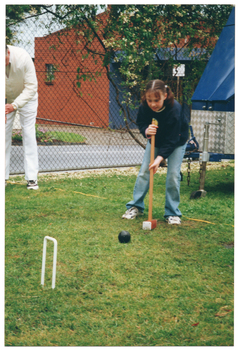COL photo of a girl holding a mallet, preparing to hit a ball through a hoop. There is an adult (partially obscured) standing nearby (at the edge of the frame). Behind the girl, there is a large covered metal trailor and a metal wire cyclone fence around the perimeter of the grassy area on which they are located. Through the fence, a brick building can be seen across the road.