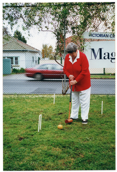 COL photo of a woman holding a mallet, preparing to hit a ball through a hoop. She is standing on a grassy area, around which there is a metal wire cyclone fence. Attached to the fence is a sign (partially obscured) with the text "VICTORIAN CR..." and "Mag...". There are two houses visible across the road, and a red sedan car travelling on the road.