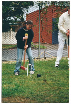 COL photo of a girl holding a mallet, preparing to hit a ball through a hoop. There is an adult (partially obscured) standing nearby (at the edge of the frame). He is also holding a mallet, as if ready to strike a ball. Behind the girl, there is a metal wire cyclone fence around the perimeter of the grassy area on which they are located. Through the fence, a house with a picket frence and a brick building can be seen across the road.
