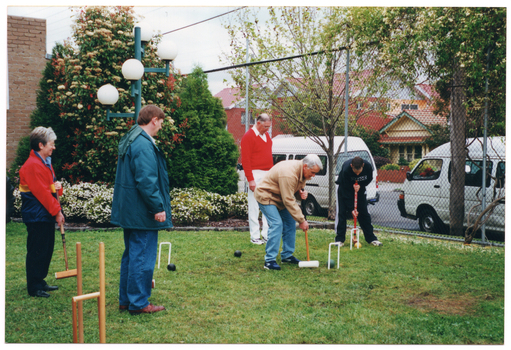 COL photo of a small group of people playing croquet on a grassy area. Some people are holding mallets, preparing to strike a ball through a hoop, while others are looking on. Behind them is a brick building and a metal wire cyclone fence surrounding the grassy area. In the corner between the building and the fence is a landscaped garden bed with some shrubs and flowering plants. There is a tall lamp in front of the garden bed. Behind the fence, two white vans can be seen parked on the road, with some houses and other buildings across the road.