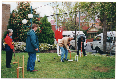COL photo of a small group of people playing croquet on a grassy area. Some people are holding mallets, preparing to strike a ball through a hoop, while others are looking on. Behind them is a brick building and a metal wire cyclone fence surrounding the grassy area. In the corner between the building and the fence is a landscaped garden bed with some shrubs and flowering plants. There is a tall lamp in front of the garden bed. Behind the fence, two white vans can be seen parked on the road, with some houses and other buildings across the road.