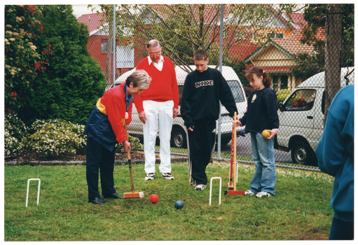 COL photo of a small group of people playing croquet on a grassy area. A woman is holding a mallet, preparing to strike a ball through a hoop. A man and two children look on. The children are also holding mallets. There is one other person (partially obscured) in the foreground, on the edge of the frame. Behind the group is a metal wire cyclone fence surrounding the grassy area. On one side, there is a landscaped garden bed with some shrubs and flowering plants. Behind the fence, two white vans can be seen parked on the road, with some houses and other buildings across the road.
