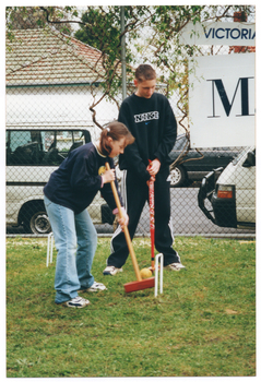 COL photo of a girl holding a mallet, preparing to strike a ball through a hoop while a boy, who is also holding a mallet, looks on. They are standing on a grassy area, fenced off by a metal wire cyclone fence. There are two signs (partially obscured) on the fence: one contains the text "VICTORI..." and the other "M...". Behind the fence, two white vans can be seen parked on the road, with some houses and another car parked across the road.