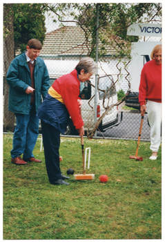 COL photo of three people playing croquet on a grassy area. One woman is holding a mallet, preparing to strike a ball through a hoop while another woman and a man look on. They are also holding mallets. Behind the group is a metal wire cyclone fence surrounding the grassy area. There are two signs (partially obscured) on the fence: one contains the text "VICTO...".  Behind the fence, a white van can be seen parked on the road, with some houses and another car parked across the road.