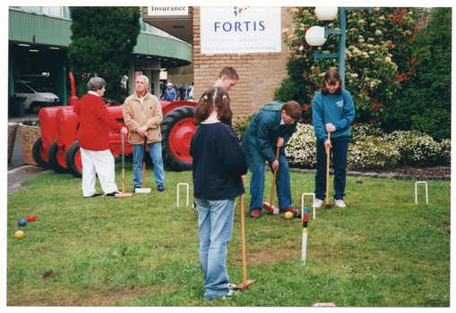 COL photo of a small group of people playing croquet on a grassy area. One adult is holding a mallet, preparing to strike a ball through a hoop, while three children look on. Another two adults are standing nearby. They are all holding mallets. Behind them, there is a red tractor next to the corner of a small brick building. There are two signs on the side of the building - one (partially obscured) contains the text "Insurance" and the other is for Fortis Group (an insurance, banking, and investment management company). There is a multistorey green building in the background with a carpark on the ground level. There are some people walking nearby.