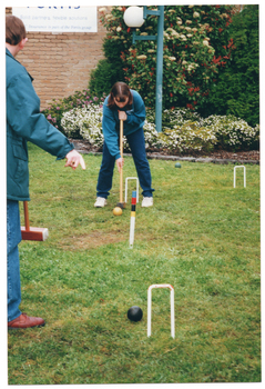 COL photo of a girl, crouching forward, holding a mallet and preparing to strike a ball. An adult is standing in the foreground (on the edge of the frame, partially obscured). They are on a grassy area. Behind them is a brick building, a tall lamp, and a garden bed with shrubs and flowering plants. There is a sign on the side of the building (partially obscured).