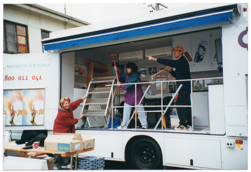 COL photo of two people, each holding a croquet mallet aloft. They are standing inside a large van, at the wide side opening. Inside the van, there are shelves, cupboards, two air conditioning units, and posters on the wall. A third person is standing at ground level below, pulling down the van's retractable metal stairs. She is standing behind a long wooden trestle table with various items on top, including two boxes and a mug. The van is parked beside a two storey building.