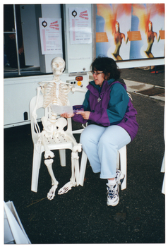 COL photo of a woman posing for a photo with a plastic skeleton. They are both seated on plastic chairs, which have been placed on the ground in front of a large van. The van has a large side door, which is in the open position. There are posters on the side of a cupboard inside the van and on the inside of the wide open door.