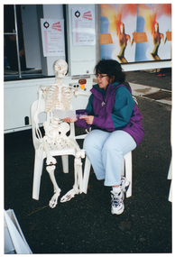 COL photo of a woman posing for a photo with a plastic skeleton. They are both seated on plastic chairs, which have been placed on the ground in front of a large van. The van has a large side door, which is in the open position. There are posters on the side of a cupboard inside the van and on the inside of the wide open door.