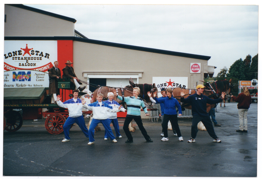 COL photo of a small group of people taking part in some Tai Chi exercises. Three people are wearing matching blue and white track suits, while the others are in casual clothes. Behind them is a wagon being pulled by two Clydesdale horses. Two people are on top of the wagon: one of them is seated and holding the reins while the other is standing behind. On the side of the wagon, there is some text (partially obscured) which reads "...MANDEL CLYDESDALES". Behind the wagon and horses, there is a building with the sign "LONESTAR STEAKHOUSE AND SALOON" and a red star logo. In front of the building is another sign for the Melbourne Markets, but it is not clear whether or not this sign is affixed to the wagon or is behind the wagon. The sign (partially obscured) says "...esale ... & ...bles", an "MM" logo with the letters colourfully illustrated with fruits, vegetables and flowers, "National Flower Centre", and "...elbourne Markets - West Melbourne". This sign refers to the wholesale Melbourne Fruit & Vegetable Market and National Flower Centre which was, at the time, located in West Melbourne. The relocation of this wholesale market to Epping was completed in 2015. See https://warequip.com.au/turnkey/melbourne-market-relocation/https://warequip.com.au/turnkey/melbourne-market-relocation/  In the background to the right of the photo, a food caravan selling doughnuts can be seen in the distance.