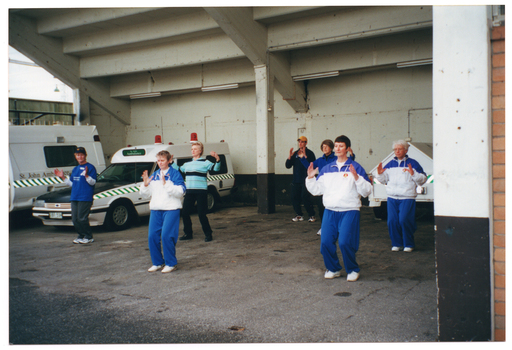 COL photo of a small group of people taking part in a Tai Chi demonstration. They are in a St John's Ambulance Australia parking bay beneath a building or stadium. There are two St John's Ambulance Australia ambulances and one trailor parked in the parking bay.