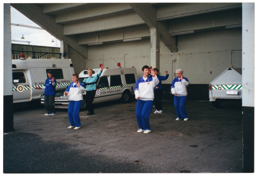 COL photo of a small group of people taking part in a Tai Chi demonstration in a St John's Ambulance Australia parking bay beneath a building or stadium. Two St John's Ambulance Australia ambulances and one trailor are parked in the parking bay.