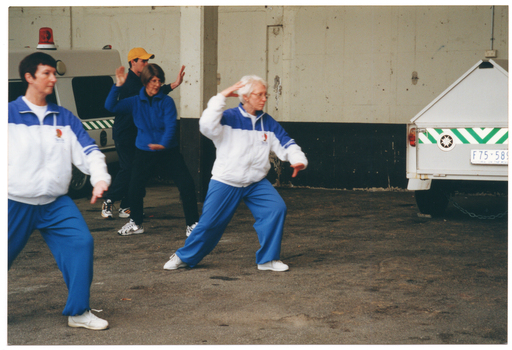 COL photo of a small group of people taking part in a Tai Chi demonstration in a St John's Ambulance Australia parking bay beneath a building or stadium. A St John's Ambulance Australia ambulance and trailor can be seen parked in the parking bay.