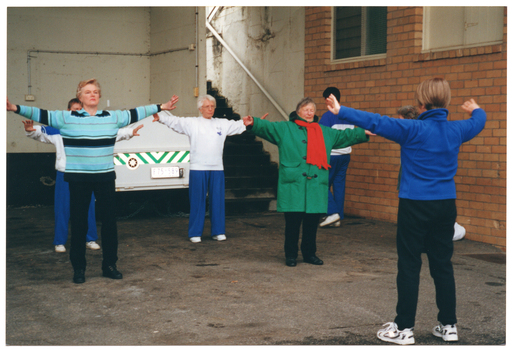 COL photo of a small group of people taking part in a Tai Chi demonstration in a St John's Ambulance Australia parking bay beneath a building or stadium. A St John's Ambulance Australia trailor is parked in the parking bay.