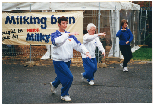 COL photo of three women taking part in a Tai Chi demonstration. Behind them is a large marquee, partially fenced off with metal wire fencing. There are straw bales and loose straw on the marquee floor. There is a large horizontal banner (partially obscured) attached to the wire fencing which reads "Milking barn brought to you by Nestlé Milky...".