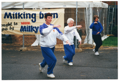 COL photo of three women taking part in a Tai Chi demonstration. Behind them is a large marquee, partially fenced off with metal wire fencing. There are straw bales and loose straw on the marquee floor. There is a large horizontal banner (partially obscured) attached to the wire fencing which reads "Milking barn brought to you by Nestlé Milky...".