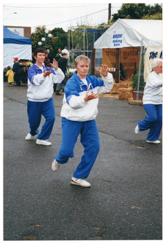 COL photo of three women taking part in a Tai Chi demonstration. Behind them are two marquees and a brick building with a sign on the exterior brick wall. One marquee is partially fenced off with metal wire fencing. There are straw bales and loose straw on the marquee floor. On the gable section of the front of the marquee, there is a sign which reads "Nestlé Milkybar Milking Barn". Next to the marquee, there is a wire fence with a large horizontal banner attached to it.