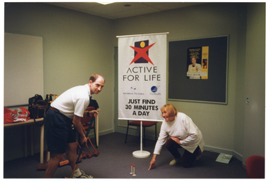 COL photo of two people demonstrating the game of croquet. A man is holding a mallet while a woman crouches down and points to a point beyond the hoop, where he needs to aim. Behind them is a large freestanding 'ACTIVE FOR LIFE' banner. The banner contains the Active for Life logo, as well as the names and logos for Arthritis Victoria and VicHealth. A sub-heading says "JUST FIND 30 MINUTES A DAY". On the wall behind them, there is a display board with a poster depicting an image of media personality, Ita Buttrose.