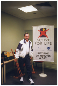 COL photo of a man holding several croquet mallets in each hand. Behind him, there is a large freestanding 'ACTIVE FOR LIFE' banner. The banner contains the Active for Life logo, as well as the names and logos for Arthritis Victoria and VicHealth. A sub-heading says "JUST FIND 30 MINUTES A DAY". There are some tables against the wall, with various croquet equipment on them.
