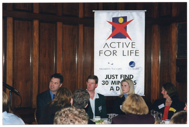 COL photo of four people, sitting at a table at a function. Three of them are looking towards the left of the frame, where there is a microphone on a stand (partially obscured). Behind them, there is a large freestanding banner titled, 'ACTIVE FOR LIFE'. The banner contains the 'Active for Life' logo, as well as the names and logos for 'Arthritis Victoria' and 'VicHealth'. A sub-heading says, "JUST FIND 30 MINUTES A DAY". Behind the banner, there is a wood-pannelled wall. There are wine glasses, some cups, a plate of sandwiches, and a glass jug on the table. In the foreground, the backs of some people's heads are evident.