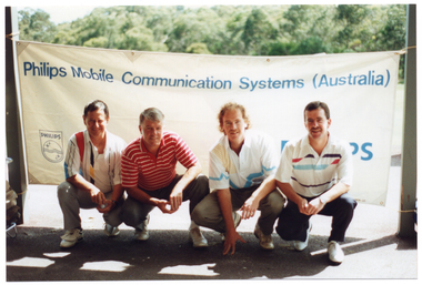 COL photo of four men crouching down in front of a large horizontal banner. The banner is attached to two vertical wooden veranda posts. The text across the top of the banner says, "Phillips Mobile Communication Systems Australia". There is a Phillips logo on the bottom left corner of the banner, and some more text (partially obscured) on the bottom right of the banner. Behind the banner, there are several large trees evident in the distance.