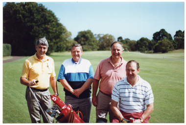 COL photo of four men on a golf course. One man is seated while the other three are standing. They are all smiling. One of the men standing has one hand on the long handle of a golf caddy, which has a golf bag and clubs on it. The man who is seated is holding a can of soft drink in one hand.