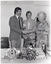 B&W photo of three people standing behind a table. A man and a woman are shaking hands while another woman stands between them. She is facing the first woman and smiling. The man is holding a cheque in his other hand. On the table in front of them, there is a small decorative 'LBA Victoria' logo on a stand, a small basket of flowers, and a small Australian flag on a stand.