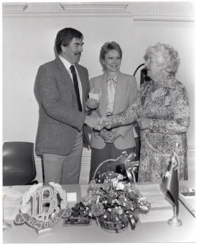 B&W photo of three people standing behind a table. A man and a woman are shaking hands while another woman stands between them. She is facing the first woman and smiling. The man is holding a cheque in his other hand. On the table in front of them, there is a small decorative 'LBA Victoria' logo on a stand, a small basket of flowers, and a small Australian flag on a stand.