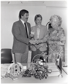 B&W photo of three people standing behind a table. A man and a woman are shaking hands while another woman stands between them. She is facing the first woman and smiling. The man is holding a cheque in his other hand. On the table in front of them, there is a small decorative 'LBA Victoria' logo on a stand, a small basket of flowers, and a small Australian flag on a stand.