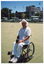COL photo of a man holding a bowling ball in his hands and looking directly at the camera. He is a wheelchair user, and is on a large grass lawn bowls court. On the edge of the court, three shelters with bench seats are evident. They each have an advertising sign on them, two of which are ANZ Bank advertisements. There are several cars parked in a car park behind the shelters, with some multistorey buildings in the background.