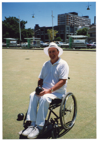 COL photo of a man holding a bowling ball in his hands and looking directly at the camera. He is a wheelchair user, and is on a large grass lawn bowls court. On the edge of the court, three shelters with bench seats are evident. They each have an advertising sign on them, two of which are ANZ Bank advertisements. There are several cars parked in a car park behind the shelters, with some multistorey buildings in the background.