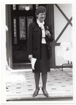 B&W photo of a woman, wearing a jacket, shirt, and skirt, standing on a tiled veranda. She is holding some papers in one hand and grimacing. There is a hand-painted and stained glass door behind her.