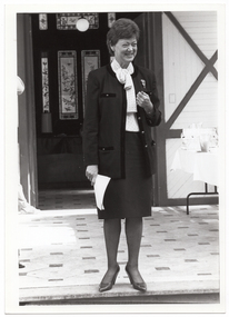 B&W photo of a woman, wearing a jacket, shirt, and skirt, standing on a tiled veranda. She is holding some papers in one hand and grimacing. There is a hand-painted and stained glass door behind her.
