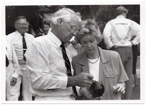 B&W medium close-up photo of a man holding a bowl (a ball used in lawn bowls) in his hands. He is speaking to a woman, and they are both looking at the ball. There are several other people in the background.