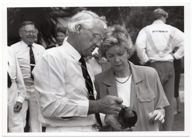 B&W medium close-up photo of a man holding a bowl (a ball used in lawn bowls) in his hands. He is speaking to a woman, and they are both looking at the ball. There are several other people in the background.