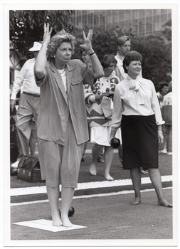 B&W photo of a group of people outdoors. There are two women in the foreground: one woman is holding her hands up around her head, as if in exasperation. The other woman, who is holding a bowl (a ball used in lawn bowls) in one hand, is looking ahead into the distance. They are both in stockinged feet. In the background, there are some people standing behind them.
