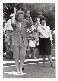 B&W photo of a group of people outdoors. There are two women in the foreground: one woman is holding her hands up around her head, as if in exasperation. The other woman, who is holding a bowl (a ball used in lawn bowls) in one hand, is looking ahead into the distance. They are both in stockinged feet. In the background, there are some people standing behind them.