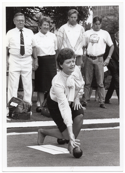 B&W photo of a woman crouching down as she bowls in her stockinged feet. There is a piece of fabric or vinyl on the ground, at her feet. Standing behind her, there are four other people looking on. There are two bowls bags at their feet.