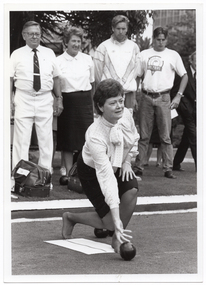B&W photo of a woman crouching down as she bowls in her stockinged feet. There is a piece of fabric or vinyl on the ground, at her feet. Standing behind her, there are four other people looking on. There are two bowls bags at their feet.