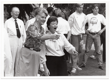 B&W photo of two women in the foreground, with several other people behind them. One woman has her arm aroun the other woman's shoulders. She is pointing to something ahead, while the other woman has her eyes and mouth open in surprise.