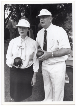 B&W photo of a man and a woman, standing outdoors. They are each holding a bowl (a ball used in lawn bowls) in their hand/s and looking ahead into the distance. There are some large trees behind them.
