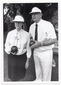 B&W photo of a man and a woman, standing outdoors. They are each holding a bowl (a ball used in lawn bowls) in their hand/s and looking ahead into the distance. There are some large trees behind them.