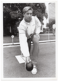 B&W photo of a man crouching down as he bowls. He is wearing a track suit jacket and pants, and runners. There is a piece of fabric or vinyl on the ground, at his feet. Behind him, there is a marker with the number "2" on it. In the background, there is a clothes rack beneath a large tree, with some jacket blazers hanging from coathangers. A house (partially obscured) is also evident, as is a multi-storey building in the distance, to the right of the frame.