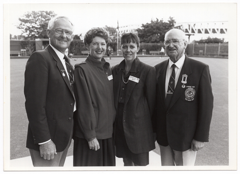 B&W photo of four people standing next to one another on a bowling green. In the background, there are a few people spread around on the green. In the distance, there are some large trees, a building, some fencing, and a bridge.